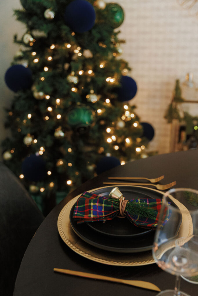 Holiday tablescape with green and navy accents, candles, and a bell napkin ring. a christmas tree is in the background.